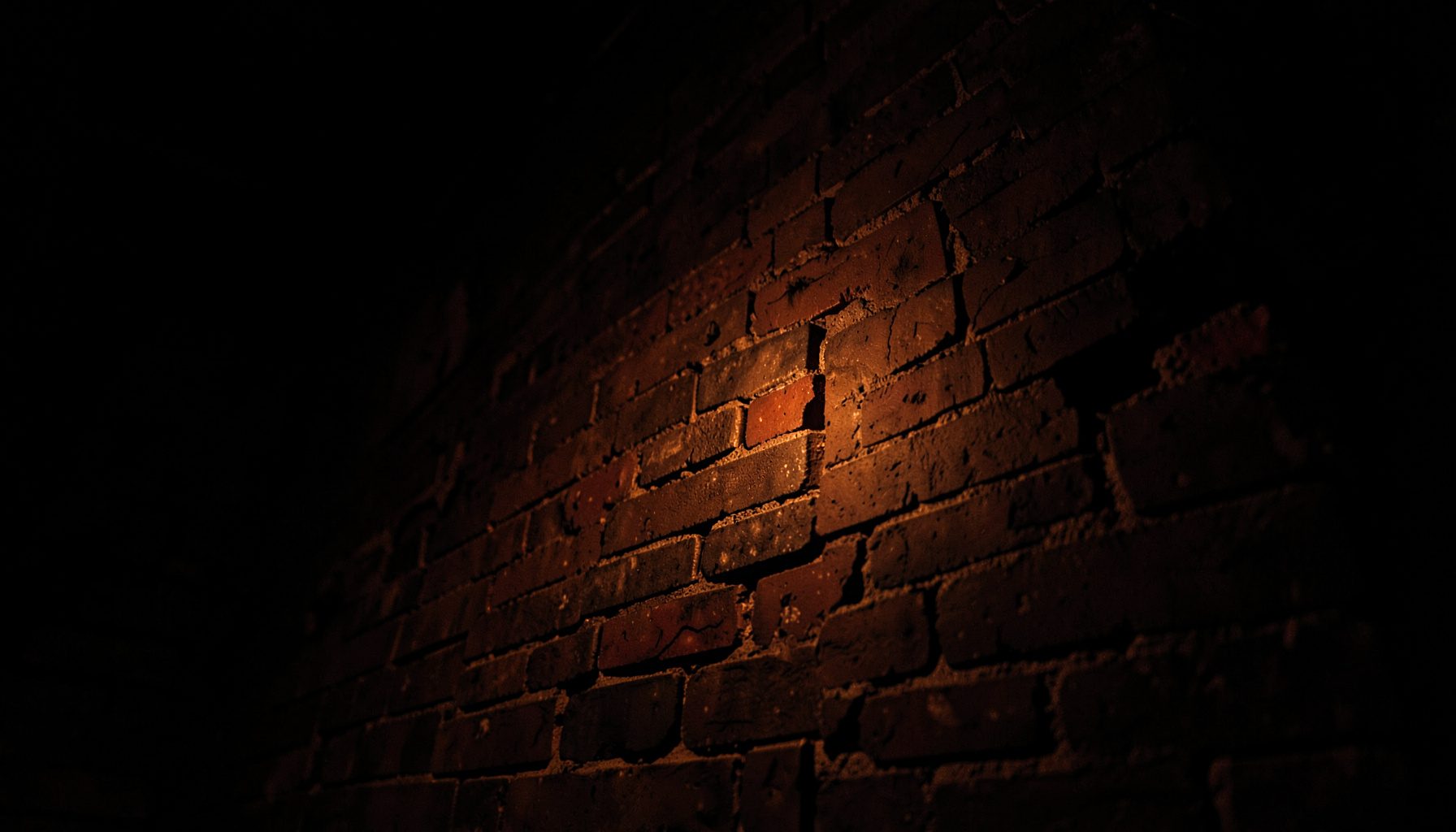 Photographie sombre en contre-plongée d'un mur de briques industrielles éclairé par une faible lumière orange, ambiance lourde et mystérieuse.
